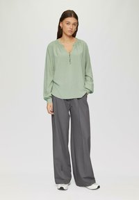 Blouse longue à manches vert clair avec un col rond et une patte de boutonnage à trois boutons, associée à un pantalon large gris et des baskets blanches.