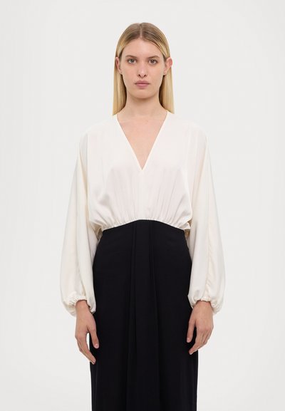 Blouse blanche à manches longues avec un décolleté en V profond et une taille froncée, associée à une longue jupe noire. Tissu lisse et fluide avec une silhouette structur�ée.