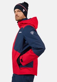 Giacca outdoor rossa e blu con cappuccio, dotata di tasche con zip, polsini regolabili e un logo sulla manica. Calda cuffia a maglia con pompon.
