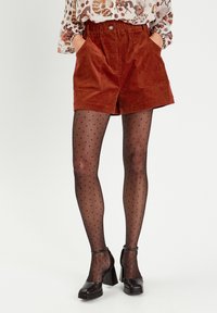 Rostfarbene Cordshorts mit zwei vorderen Taschen, kombiniert mit schwarzen Pünktchenstrumpfhosen und klobigen schwarzen Absätzen, die einen stylischen, geschichteten Look präsentieren.