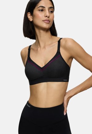 Femme portant un brassière de sport noire texturée avec des bretelles fines et des accents violets, associée à un legging noir taille haute.