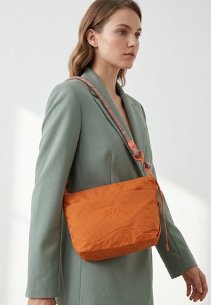 Femme portant un blazer vert sauge porte un sac bandoulière orange avec une sangle à motif sur l'épaule.