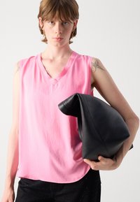 Roze mouwloze top met een v-hals, gemaakt van een gladde stof. Het model houdt een grote zwarte leren tas vast met een zachte textuur.