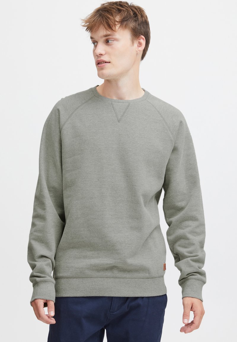 Sudadera gris claro con mangas raglán, cuello y puños de canalé, y una etiqueta de logo discreta en el dobladillo. Tejido suave y texturizado.