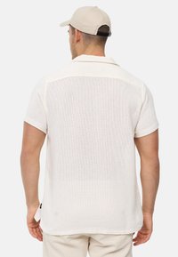 Weißes Poloshirt mit kurzen Ärmeln aus strukturiertem Strick, mit klassischem Kragen und geradem Saum, von hinten gezeigt.