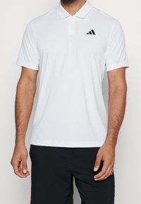 Polo blanc en tissu respirant, avec un col classique et des manches courtes. Comprend un logo Adidas noir sur la poitrine gauche.