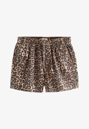 Vrouw met krullend haar staat blootsvoets op het zand, draagt een wit shirt met een zonafbeelding en shorts met luipaardprint voor een grote rots.