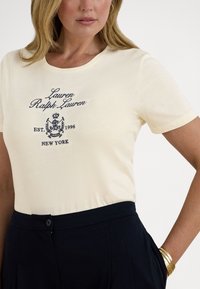 Κοντομάνικη κρεμ μπλούζα με κεντημένο με μπλε σκούρο κείμενο και λογότυπο. Φοριέται με μπλε σκούρο παντελόνι και βραχιόλια σε χρυσό τόνο. Υφή υφάσματος λεία.