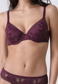 Soutien-gorge in pizzo mauve con ferretto, motivo floreale, spalline regolabili e bordi ondulati. Presenta un gancio centrale tra le coppe per la regolazione.