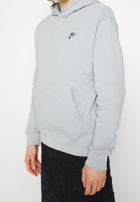 Světlý šedý mikina s kapucí vyrobená z měkké tkaniny, se středovou klokaní kapsou a malým černým logem Nike na hrudi.
