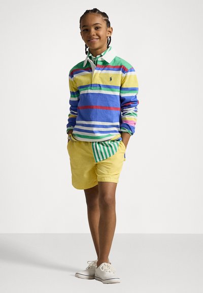 Flerfarget stripete langermet polo skjorte med blå, grønn, gul og rød stripe; kombinert med gule shorts og hvite sko.