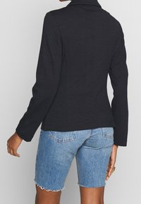 Veste noire à manches longues en tissu texturé, coupe ajustée, associée à un short en denim bleu clair aux bords effilochés.