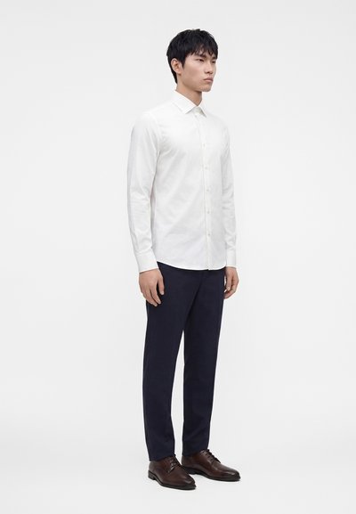Mężczyzna stojący, ubrany w białą koszulę zapinaną na guziki, ciemne spodnie i brązowe eleganckie buty, na jednolitym białym tle.