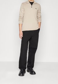 Pull beige à fermeture éclair avec logo, assorti à un pantalon noir ample et des chaussures noires. Tissu lisse, design décontracté, accents minimalistes.