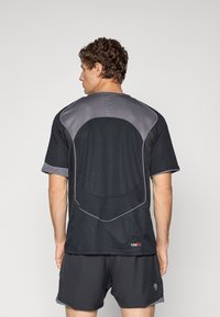 Man with curly hair wearing black and gray sportswear, viewed from the back, standing against a plain light background.