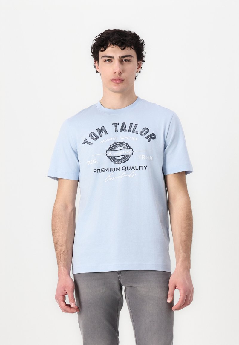 Tricou din bumbac de culoare bleu deschis, cu un imprimeu grafic întunecat care conține textul "TOM TAILOR" și un logo circular. Decupaj la gât și mâneci scurte.
