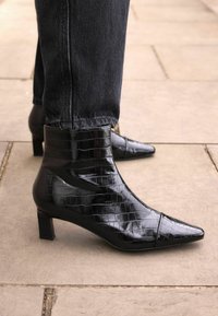 Mustad mustnahkse krokodillimustriga ankle boots, terava ninaga ja tüsedate kontsadega, koos tumedate denim-pükstega.