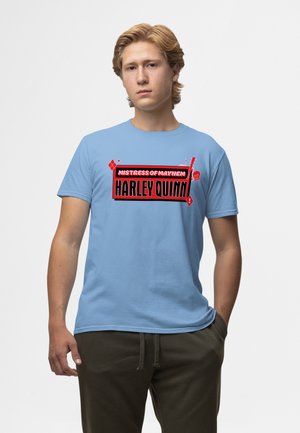 Giovane uomo con capelli chiari che indossa una T-shirt azzurro chiaro con la grafica rossa e nera "Mistress of Mayhem Harley Quinn", in piedi con una mano in tasca.