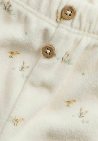 Luddigt vitt tyg med små, gula kycklingillustrationer och gröna blommiga inslag. Två träknappar synliga nära midjan.