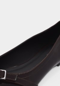 Σκούρο καφέ δερμάτινο επίπεδο παπούτσι με μία μικρή ασημένια αγκράφα κοντά στη μύτη, που δείχνει την εσωτερική σόλα και τις ραφές στο πλάι.