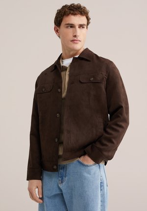 Man draagt een bruin jasje met knopen over een beige trui en wit overhemd, met lichtblauwe spijkerbroek, staand met één hand in zijn zak.