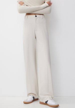 Person iført cremefarvede bukser med vide ben, off-white ribbet langærmet top og hvide sneakers med sorte striber og brune såler.