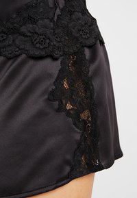 Melnas satīna šorti ar mežģīņu apdari, kurai ir ziedu raksti augšā un sānos, piešķirot dizainam tekstūru un detaļas.