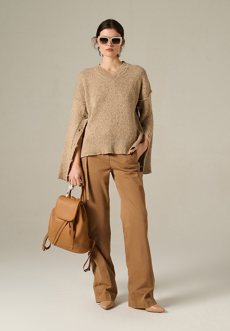 Maglione beige a maglia con maniche larghe, abbinato a pantaloni color cammello. Tiene uno zaino in pelle marrone e indossa occhiali da sole bianchi.