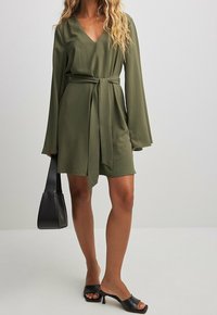 Frau in einem olivgrünen langärmligen Kleid mit Gürtel, die eine schwarze Handtasche hält und schwarze offene Heels trägt, steht auf einem weißen Boden.