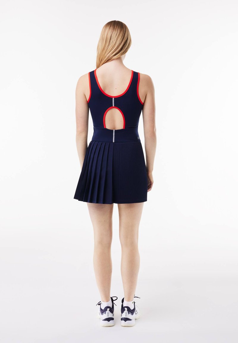 Marineblauwe jurk met een geplooide rok, met een uitgesneden achterkant en rode accenten. Gemaakt van elastisch materiaal, gecombineerd met witte sportschoenen.