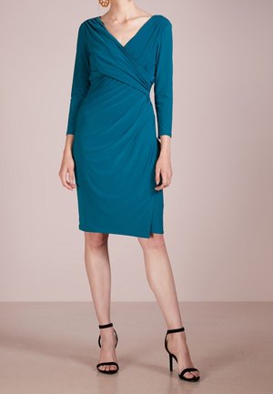 Sukienka w kolorze morskim wykonana z miękkiego materiału, z dekoltem w serek, długimi rękawami, drapowaniem i bocznym rozcięciem, zestawiona z czarnymi wysokimi sandałami na paskach.