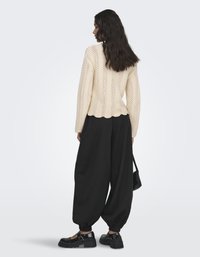 Femme aux longs cheveux bruns portant un pull crème texturé, un pantalon noir ample retroussé et des chaussures noires épaisses, tenant un petit sac noir.