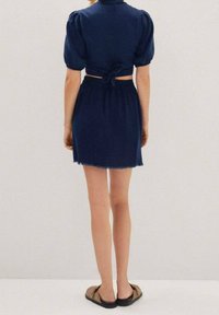 Kort marineblå kjole med pufærmer, indsnøret talje og en detalje med bindebånd bagpå. Frynset kant og kombineret med brune slip-on sko.