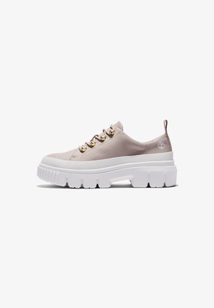 Jasnobeżowy niski sneaker z grubą białą teksturowaną podeszwą, złotymi oczkami, materiałową pętlą na pięcie i małym logo drzewa w pobliżu kostki.