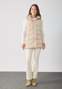 Gilet matelassé beige avec un col montant, fermeture à pression sur le devant et poches latérales. Porté par-dessus un haut à manches longues blanc et associé à un pantalon crème.