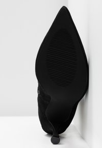 Svart mocka högklackad sko med spetsig tå, som har en texturerad gummisula och en elegant, minimalistisk design.