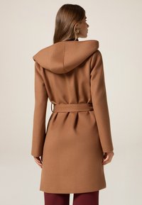 Bruine gewatteerde jas met een ceintuur in de taille, naadloos ontwerp en een stevige stof. Heeft een lichte plooi detail aan de achterkant.