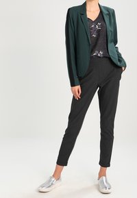Blazer verde scuro sopra un top nero a fantasia, abbinato a pantaloni neri a sigaretta e scarpe slip-on argento. Tessuto morbido e vestibilità su misura.