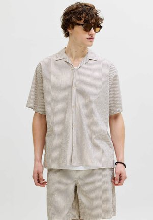 Muž v béžovej a bielej vertikálne pruhovanej košeli s krátkym rukávom na gombíky a zodpovedajúcich krátkych nohaviciach, so hnedými slnečnými okuliarmi a čiernym náramkom.