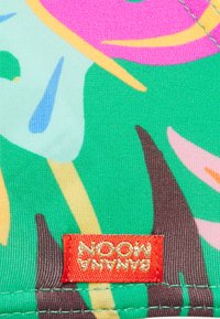 Tecido com um fundo verde vibrante, apresentando formas abstratas em rosa, azul e amarelo. Uma etiqueta vermelha com "BANANA MOON" é visível.
