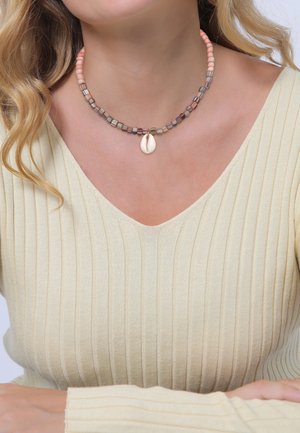 Femme portant un collier de perles avec un pendentif central en coquillage cauri blanc et un pull crème à col en V côtelé.