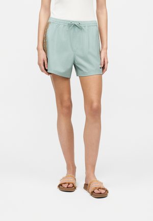 Femeia poartă pantaloni scurți verde deschis cu șnur, cu panouri laterale maro deschis cu model și sandale bej cu barete multicolor țesute.