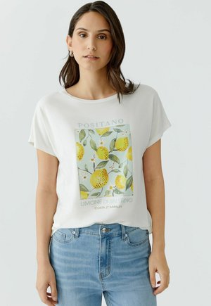 Frau trägt weißes T-Shirt mit Zitronen- und Blätterdruck und dem Text „Positano Limone di Salerno Costa d'Amalfi“ kombiniert mit blauen Jeans.