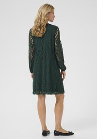 Rochie din dantelă verde închis, cu mâneci lungi, model floral și tiv evazat, asortată cu balerini negri care au un detaliu mic în formă de fundiță.