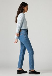 Camicia azzurro chiaro a maniche lunghe abbinata a jeans blu aderenti con una toppa rossa sul taschino posteriore e mocassini neri.