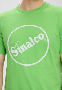 Groene katoenen T-shirt met een groot wit rond logo met het woord "Sinalco" in vette letters gecentreerd op de voorkant. Korte mouwen.