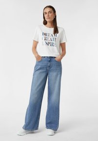 Weißes Baumwoll-T-Shirt mit grafischem Druck; kombiniert mit hellblauen, weit geschnittenen Jeans; Model trägt weiße Sneakers.