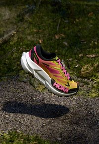 Botte de course jaune vif et rose avec une épaisse semelle blanche, flottant au-dessus d'un chemin de gravier entouré de feuillage vert.