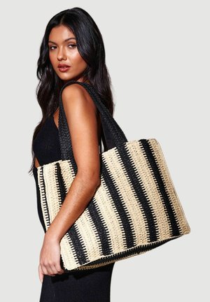 Femme aux longs cheveux foncés portant sur l'épaule un grand sac cabas tissé à rayures verticales noires et beiges.