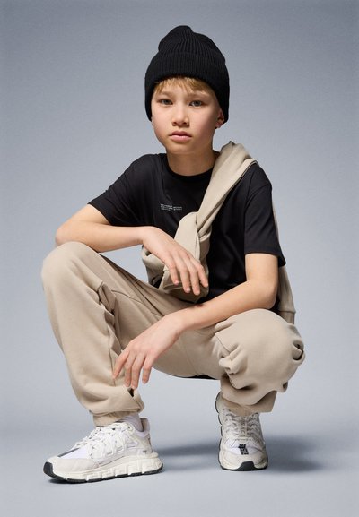 Svart t-shirt med text, beige sweatpants med elastiska muddar, vita sneakers med grå accenter och en svart stickad mössa.